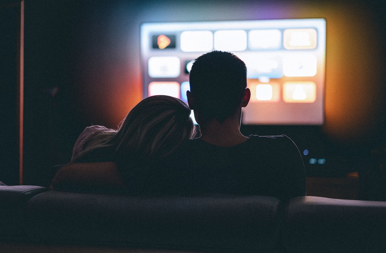 A couple sitting together on a couch, watching a television screen in a cozy living room setting.